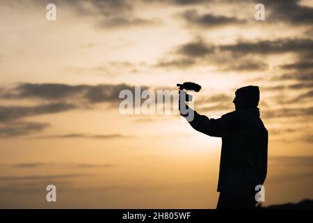 Seitenansicht Silhouette von unkenntlich junge Bloggerin in Hut Dreharbeiten Video auf Digitalkamera gegen Sonnenuntergang Himmel auf dem Land Stockfoto