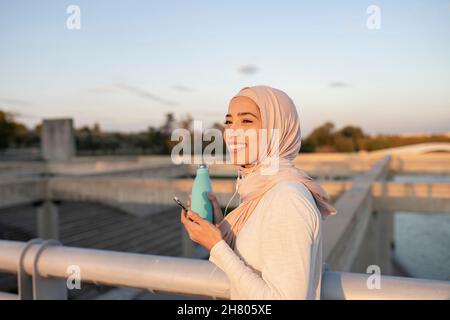 Seitenansicht einer lächelnden arabischen Frau im Hijab, die mit einer Flasche Wasser steht und Musik in Kopfhörern auf der Brücke genießt Stockfoto