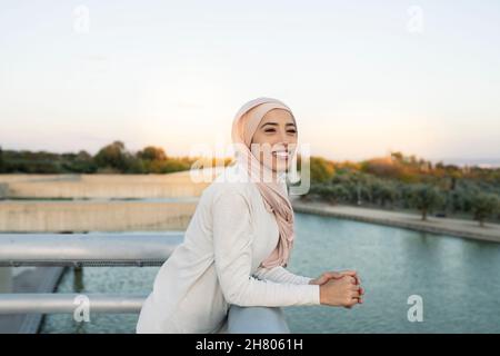 Fröhliche arabische Frau in Hijab, die mit einem Lächeln in die Ferne blickt, während sie sich am Wasser in der Nähe des plätschernden Flusses auf Metallgeländer stützt Stockfoto