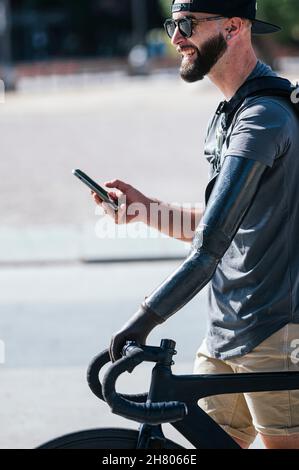 Seitenansicht eines fröhlichen bärtigen Mannes mit künstlichem Arm, der mit dem Fahrrad auf der Straße unterwegs ist, während er am sonnigen Tag auf dem Mobiltelefon stöbert Stockfoto