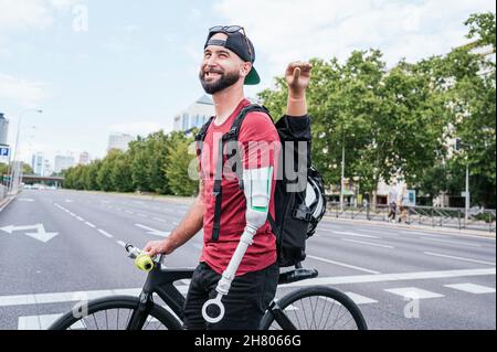 Seitenansicht eines fröhlichen männlichen Hipsters mit Armprothese, der mit dem Fahrrad auf dem Crosswalk in der Stadt läuft Stockfoto