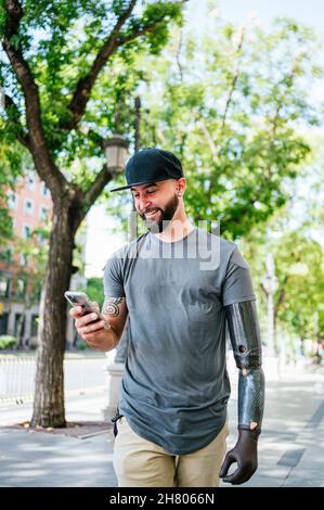 Positiver männlicher Hipster mit Armprothese-Messaging in den sozialen Medien über Handy, während er im Sommer auf der Stadtstraße spazierengeht Stockfoto
