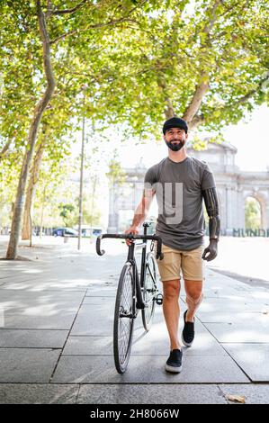 Lächelnder männlicher Hipster mit Armprothese, der mit dem Fahrrad auf dem Bürgersteig in der Stadt unterwegs ist und dabei die Kamera anschaut Stockfoto