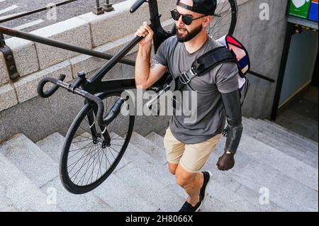 Hoher Winkel des männlichen Hipsters mit Prothese des Armes, der Fahrrad hält und Steinstufen in der Stadt hinaufgeht Stockfoto