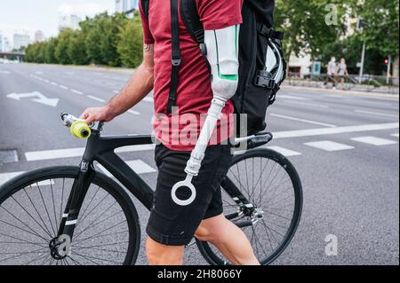 Seitenansicht des abgeschnittenen, nicht erkennbaren männlichen Hipsters mit Armprothese, der auf dem Fußgängerüberweg in der Stadt mit dem Fahrrad unterwegs ist Stockfoto