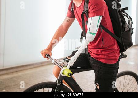 Beschnittene Seitenansicht des nicht erkennbaren bärtigen männlichen Hipsters mit künstlichem Arm, der im Sommer in der Stadt auf der Straße Fahrrad fährt Stockfoto