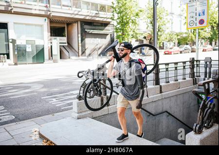 Hoher Winkel des männlichen Hipsters mit Prothese des Armes, der Fahrrad hält und Steinstufen in der Stadt hinaufgeht Stockfoto