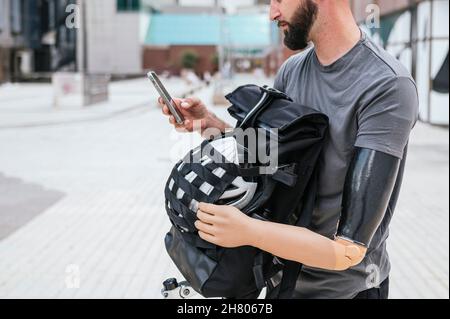 Beschnittener, nicht erkennbarer männlicher Hipster mit Armprothese Messaging in sozialen Medien über Handy während er im Sommer mit Rucksack A in der Stadtstraße spazierenging Stockfoto