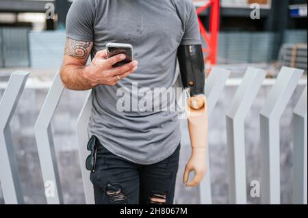 Kurzgeschnittenes, nicht erkennbares männliches Hipster mit Armprothese-Messaging in sozialen Medien über Handy, während es im Sommer auf der Stadtstraße spazierenging Stockfoto