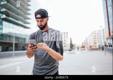 Positiver männlicher Hipster mit Armprothese-Messaging in den sozialen Medien über Handy, während er im Sommer auf der Stadtstraße spazierengeht Stockfoto
