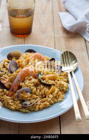 Teller mit leckerer frischer Paella mit Muscheln und Garnelen auf Holztisch mit Glas erfrischendem Getränk und Besteck serviert Stockfoto