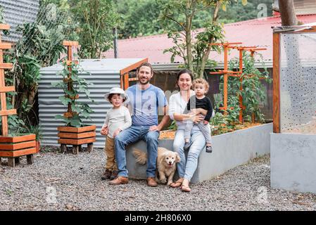 Ganzer Körper von glücklichen Eltern mit niedlichen Söhnen sitzen auf Gartenbett mit Golden Retriever Welpen auf dem Land Blick auf die Kamera Stockfoto