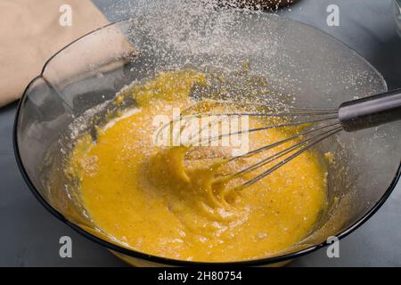 Mehl, das in eine Glasschüssel mit rohem, geschlagenem Eigelb gegossen wird, und Schneebesen, der während des Kochvorgangs auf den Tisch in der Küche gelegt wird Stockfoto