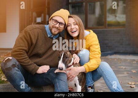 Inhalt Paar schaut auf die Kamera, die im Herbst auf einer Treppe mit Bull Terrier in der Nähe der Hausglastür sitzt Stockfoto