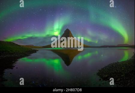 Farbenfrohe grüne Polarlichter, die am Sternenhimmel über einem ruhigen See mit felsiger Formation in wilder Natur nachts leuchten Stockfoto