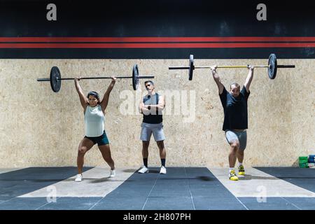 Ganzkörpergruppe starker hispanischer Sportler, die während des Gewichtheben-Trainings gemeinsam schwere Langhanteln in der Nähe der Wand in einer leichten Turnhalle heben Stockfoto