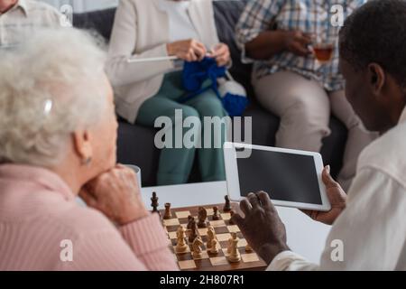 Digitales Tablet in den Händen eines afroamerikanischen Mannes in der Nähe des Schachbretts und von Freunden im Pflegeheim Stockfoto