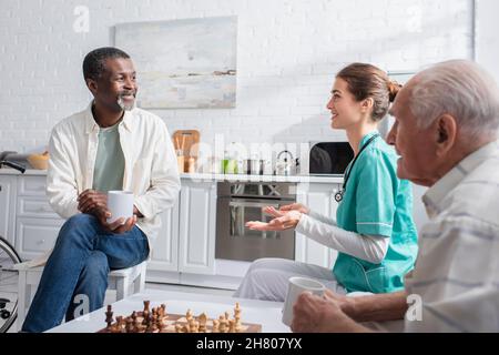 Krankenschwester, die im Pflegeheim mit interracial Patienten mit Tassen in der Nähe des Schachbretts spricht Stockfoto