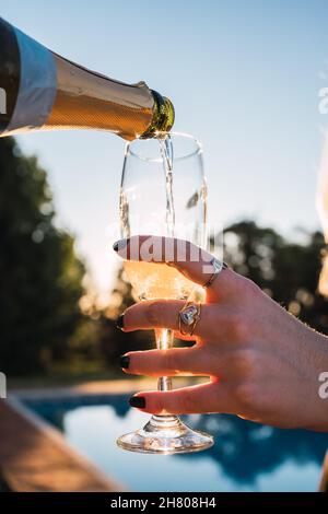 Eine anonyme Person, die am Abend vor dem unscharfen Hintergrund des Pools Champagner aus der Flasche in ein Glas gegossen hat Stockfoto
