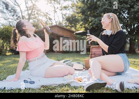 Ganzer Körper von fröhlichen jungen Freundinnen in lässigen Kleidern lachen beim Biertrinken auf der Decke sitzen während Picknick in der Natur am sonnigen Sommer d Stockfoto