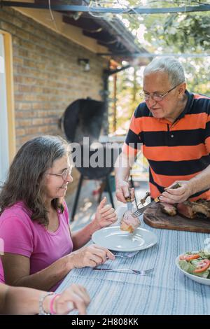 Aufmerksamer älterer Mann in legerer Kleidung, der während des Mittagessens mit der Familie auf der Terrasse des Hauses auf dem Land Gerichte serviert, die am Tisch stehen Stockfoto