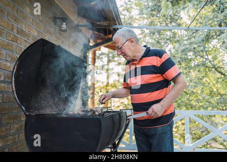Positiver älterer Mann in legerer Kleidung und Brillen, der während des Urlaubs auf dem Land Fleisch auf dem Grill auf der Terrasse des Backsteinhauses braten Stockfoto