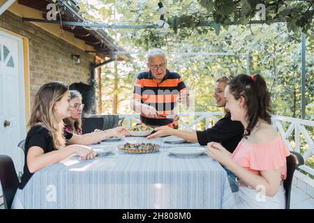 Aufmerksamer älterer Mann in legerer Kleidung, der während des Mittagessens mit der Familie auf der Terrasse des Hauses auf dem Land Gerichte serviert, die am Tisch stehen Stockfoto