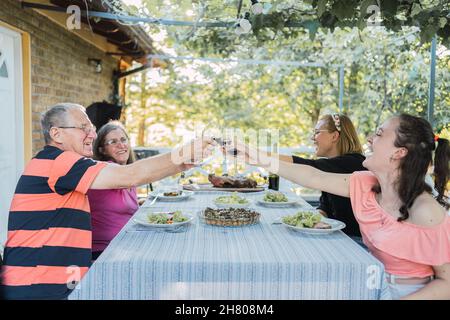 Seitenansicht einer fröhlichen Familie mit älteren Ehepartnern und jungen Töchtern, die beim Familienessen auf der Terrasse in Count lächeln und auf ein Glas Rotwein klicken Stockfoto