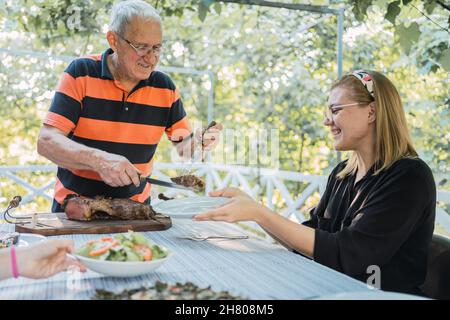 Aufmerksamer älterer Mann in legerer Kleidung, der während des Mittagessens mit der Familie auf der Terrasse des Hauses auf dem Land Gerichte serviert, die am Tisch stehen Stockfoto