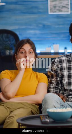 Junge interracial paar Fernsehen essen Popcorn im Wohnzimmer. Verheiratete, multiethnische Menschen, die die Kamera anschauten und einen Imbiss genossen und entspannende Aktivitäten genossen. Liebhaber gemischter Rennen Stockfoto