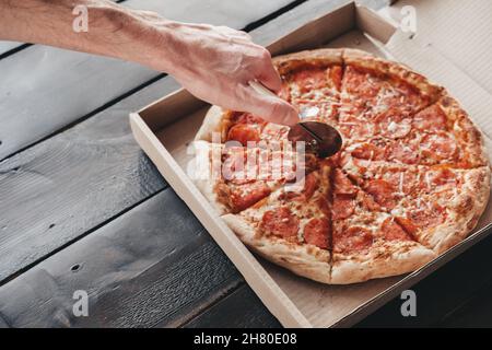 Männliche Hand schneidet Pepperoni Pizza mit einem runden Messer auf einem dunklen Holzhintergrund. Leckere und frische Pizza in einer Schachtel mit runden Würstchenstücken Stockfoto