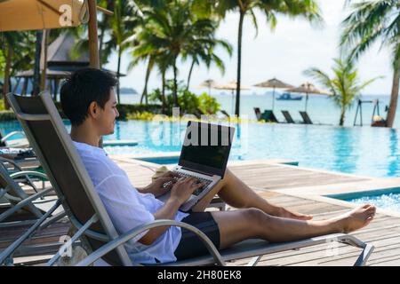 Der asiatische Mann verbrachte seinen Sommerurlaub mit der Arbeit an seinem Laptop in einem Stuhl in der Nähe des Swimmingpools im Resorthotel in der Nähe des Meeres. Stockfoto