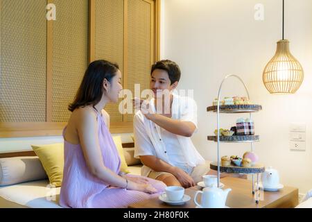 Asiatische Paare genießen einen romantischen Urlaub. Bei einem Nachmittagstee, während ein Mann seiner Freundin in einem luxuriösen Hotelzimmer Kuchen zufüttert. Stockfoto