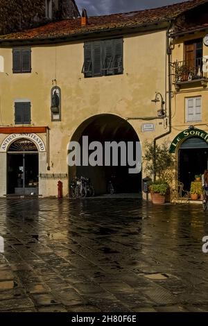 Am frühen Abend Reflexionen nach heftigem Regen auf der Piazza dell’Anfiteatro, dem öffentlichen Freigelände, das 1830 auf dem Gelände des elliptischen römischen Amphitheaters mit 10.000 Sitzplätzen in Lucca, Toskana, Italien, eingerichtet wurde. Vier gewölbte Tunnel durch die ockerfarbenen mittelalterlichen Wohnhäuser, die auf den römischen Ruinen errichtet wurden, erlauben den Blick auf ihre konvexen Rückwände, mit Ziegelsteinen und Steinschutt, die massive römische Bögen füllen, die einst die Kavea oder die Sitzmöbel unterstützten. Stockfoto