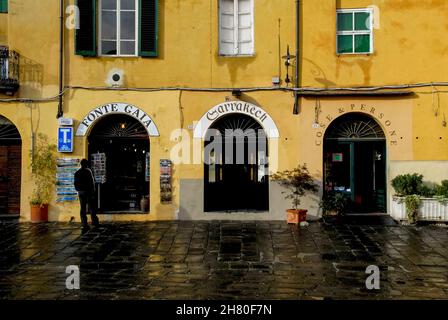 Türen zu Geschäften und Restaurants auf der Piazza dell’Anfiteatro in Lucca, Toskana, Italien, der öffentlichen Freifläche, die 1830 auf dem Gelände des elliptischen römischen Amphitheaters der Stadt mit 10.000 Sitzplätzen eingerichtet wurde. Vier gewölbte Tunnel durch die ockerfarbenen mittelalterlichen Wohnhäuser, die auf den römischen Ruinen errichtet wurden, erlauben den Blick auf ihre konvexen Rückwände, mit Ziegelsteinen und Steinschutt, die massive römische Bögen füllen, die einst die Kavea oder die Sitzmöbel unterstützten. Stockfoto