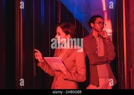 Ingenieur mit digitalem Tablet, der im Rechenzentrum in der Nähe eines nachdenklichen afrikanisch-amerikanischen Kollegen im Neonlicht arbeitet Stockfoto