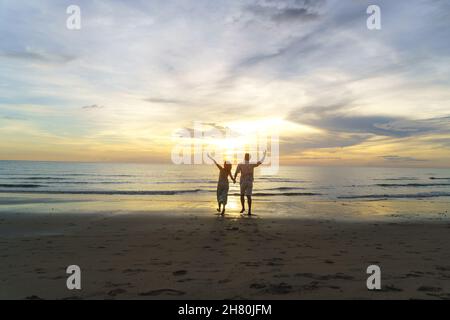Asiatische Frau Reisende halten Mann die Hand und schauen schönen Sonnenuntergang am Strand, Paar im Urlaub im Sommer und Freiheit Konzept Stockfoto