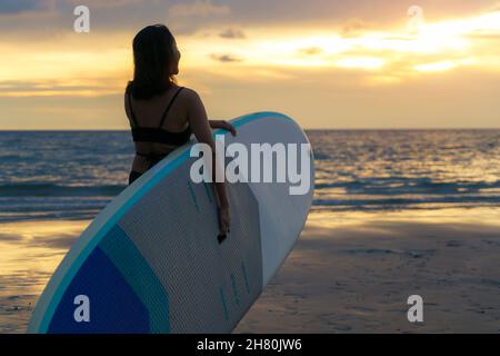 Asiatische Frau hält ein Brett und paddeln und gehen am Strand. Im Hintergrund das Meer und der Sonnenuntergang. Rückansicht. Surfen im Sommer. Stockfoto