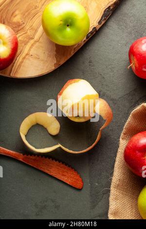 Äpfel verschiedener Sorten sind auf dunklem Stein und Holzhintergrund zu sehen. Standbild mit halb geschältem Apfel und Holzmesser. Vielseitiger Concep Stockfoto