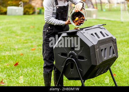 Eine Frau kippt einen kleinen Mülleimer mit Küchenabfällen in einen umstürzenden Komposter im Garten des Hinterhofs. Diese Kunststoffeinheiten mit Metallbeinen können eine drehen Stockfoto