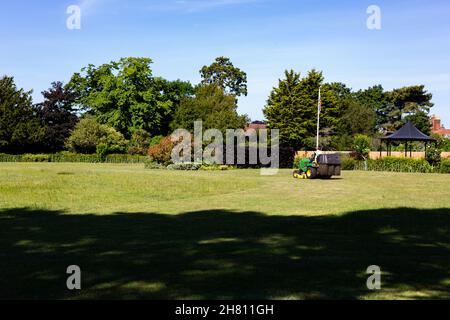 Ein ratsarbeiter, der an einem heißen Sommertag auf einem Rasenmäher in einem öffentlichen Park sitzt und das Gras schneidet Stockfoto