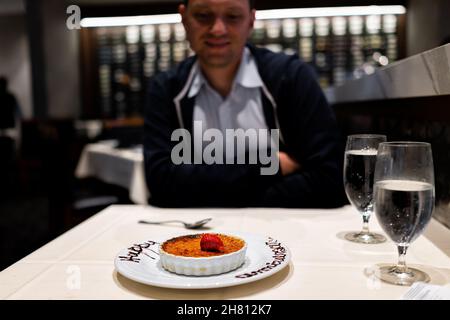 Junger glücklicher Mann, der zum Abendessen im Restaurant saß und das Essen auf dem Date mit französischem Creme-Brulee-Dessert und einem Schild zum Happy Anniversary ansah Stockfoto