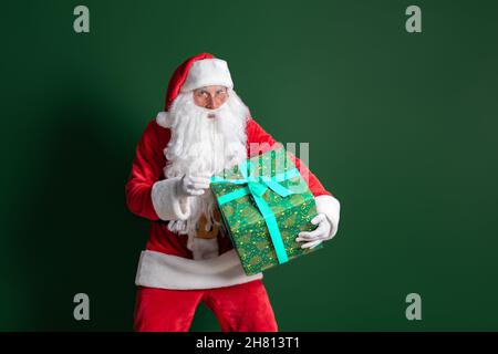 Der Weihnachtsmann mit langem Bart hält eine riesige grüne Geschenkbox auf grünem Hintergrund, während er auf die Kamera schaut Stockfoto