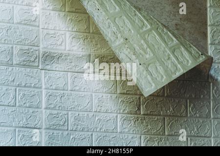 Die Tapete beginnt sich abzulösen. Die Tapete aus alten weißen Ziegeln schälte sich von der Wand im Raum, weil es feucht war oder sich nicht gut klebte. Reparieren Sie A Stockfoto