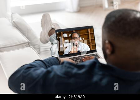 Afroamerikanischer Mann, der Online-Sprachkurse besucht, Online-Lehrer auf dem Laptop-Bildschirm. Zwei männliche, multirassische Angestellte, die online sprechen, geschäftliche Aufgaben aus der Ferne besprechen, Freunde, die online chatten Stockfoto