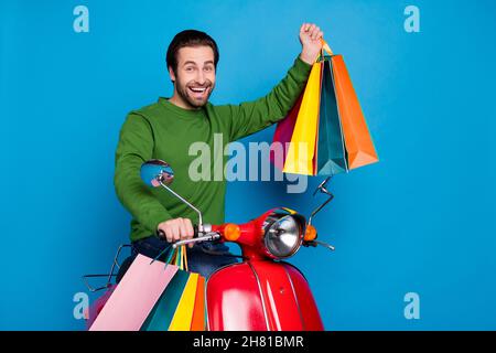Foto von positiv lächelnden jungen Mann reiten Einkaufen in roten Moped heben Fäuste halten Taschen isoliert auf blauem Hintergrund Stockfoto