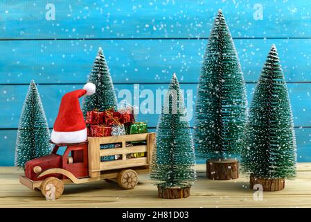 Weihnachtsgeschenke auf einem To Truck mit Weihnachtsmann-Hut neben Tannenbäumen, während es auf einem Holztisch schneit. Stockfoto