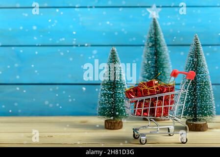 Weihnachtseinkaufskonzept. Weihnachtsgeschenke in einem Einkaufswagen neben Tannenbäumen, während es auf einem Holztisch schneit. Stockfoto