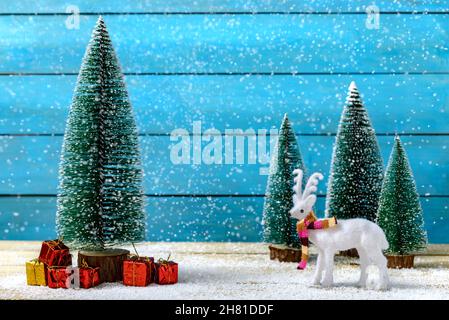 Miniatur-Rentiere, die in der Nähe von Tannen stehen, und Weihnachtsgeschenke unter fallendem Schnee auf einem Holztisch. Konzept der Feier von Weihnachten und Neujahr. Stockfoto