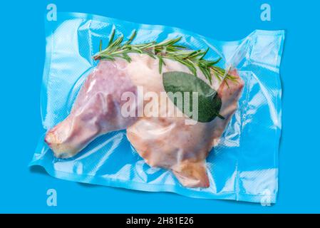 Ganze Hähnchenschenkel mit Rosmarin und Salbei in vakuumverpackter Verpackung, abgedichtet für Sous-Vide-Kochen isoliert auf blauem Hintergrund Stockfoto